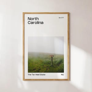 Puede incluir: Una impresión enmarcada con un diseño minimalista que presenta el texto "North Carolina" y "The Tar Heel State" con una imagen de paisaje brumoso de un poste indicador de madera en un campo. El texto "Est. 1777" y "NC" también se incluyen.