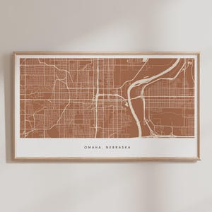 Puede incluir: Una impresión enmarcada de un mapa de Omaha, Nebraska. El mapa presenta una detallada cuadrícula de calles y un río en un esquema de color terracota y crema. El texto "OMAHA, NEBRASKA" está impreso en la parte inferior del mapa.