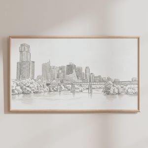 May include: A framed pencil sketch of a city skyline, featuring tall buildings, bridges, and a river. The artwork is in grayscale, with detailed shading creating depth and texture. The frame is a light wood color.