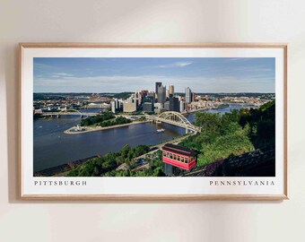 Könnte beinhalten: Ein Panoramablick auf die Skyline von Pittsburgh mit einer roten Seilbahn im Vordergrund. Die Stadt liegt an einem Fluss, über den eine Brücke führt. Der Text "PITTSBURGH" und "PENNSYLVANIA" befindet sich am unteren Rand des Bildes.