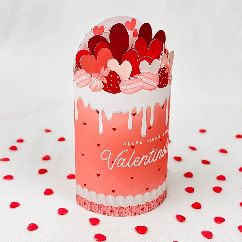 May include: A cylindrical Valentine's Day-themed gift box with a red and white color scheme. The box is decorated with hearts, macarons, and strawberries. The text "Alles Liebe zum Valentinstag" and "Valentinstag" is visible on the box.