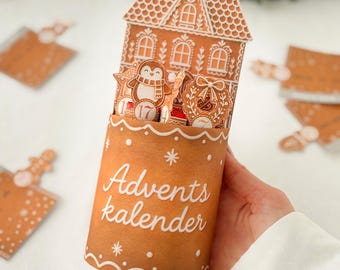 May include: A cylindrical cardboard advent calendar featuring a gingerbread house design with white icing details. The front displays the text Advents kalender. Inside the house structure are small cutouts of a penguin and a wreath.