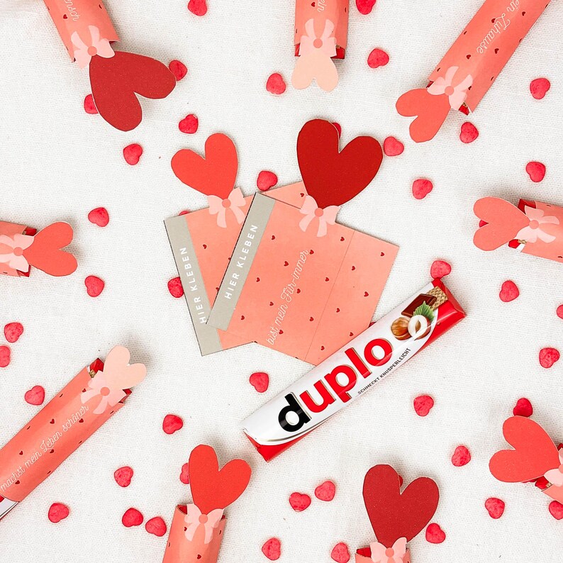 May include: A Valentine's Day-themed arrangement featuring red heart-shaped decorations, pink gift cards with German text, and a Duplo chocolate bar. The scene is set on a white surface scattered with small red heart confetti.