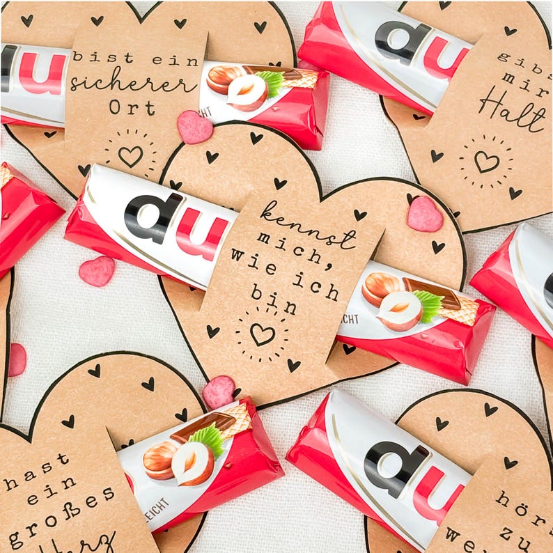 May include: An overhead shot of heart-shaped cards and chocolate bars. The cards are brown with black heart accents and handwritten text. The chocolate bars have red and white packaging. Small pink heart-shaped candies are scattered around.