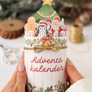 May include: A cylindrical advent calendar with vertical pink stripes and the text Advents kalender in red. The top features cutouts of a Christmas tree, a gingerbread man, a penguin, and Santa Claus, all tied with a gold metallic string bow.
