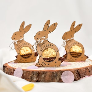 May include: Three brown cardboard Easter bunny-shaped gift boxes, each holding a gold-wrapped chocolate. The boxes are tied with white string and sit on a wooden slice. The text "Frohe Ostern!" is printed on one of the boxes.