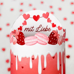 May include: A white and red decorative container with a heart and strawberry design. The container has a dripping effect with small hearts. The top features the words "mit Liebe", two pink macarons, and red strawberries.