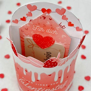 May include: A Valentine's Day gift box with a pink and white design, featuring a card that says "F&uuml;r dich" and a red heart. The box has a decorative edge and the words "Alles Liebe zum Valentinstag" are printed on the side.