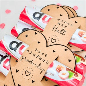 May include: A close-up of several Kinder Bueno chocolate bars wrapped in red and white packaging. Heart-shaped tags with handwritten text and small heart illustrations are placed around the bars. The text on the tags reads "gibst mir Halt" and "bist einfach gro&szlig;artig".