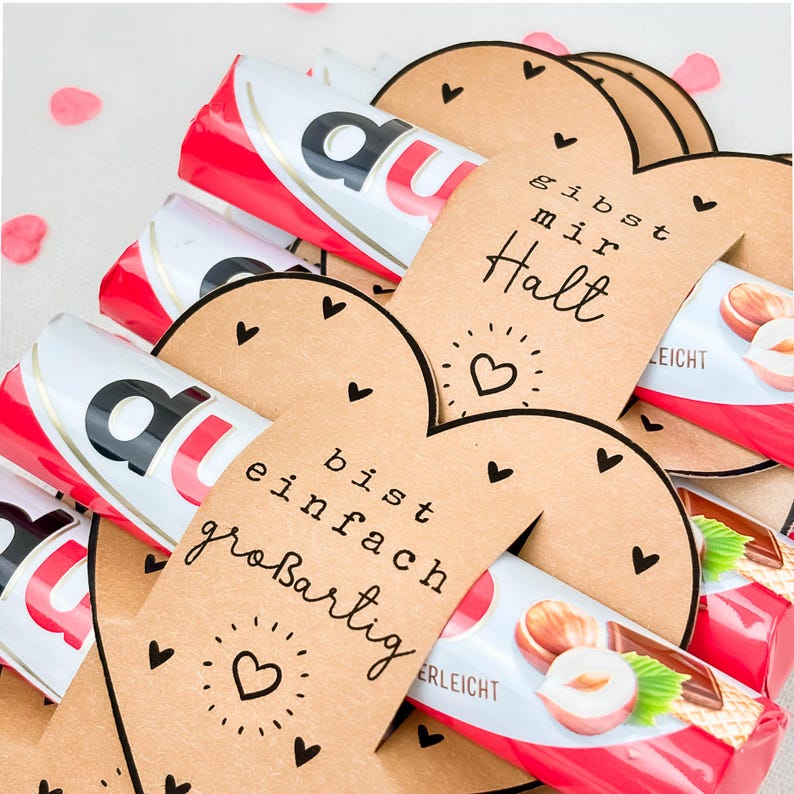 May include: A close-up of several Kinder Bueno chocolate bars wrapped in red and white packaging. Heart-shaped tags with handwritten text and small heart illustrations are placed around the bars. The text on the tags reads "gibst mir Halt" and "bist einfach gro&szlig;artig".