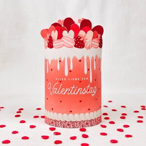 May include: A cylindrical Valentine's Day gift box with a red and pink design. The box features a dripping white frosting effect, heart accents, and the text "ALLES LIEBE ZUM Valentinstag". The top is decorated with heart-shaped cutouts and macarons.