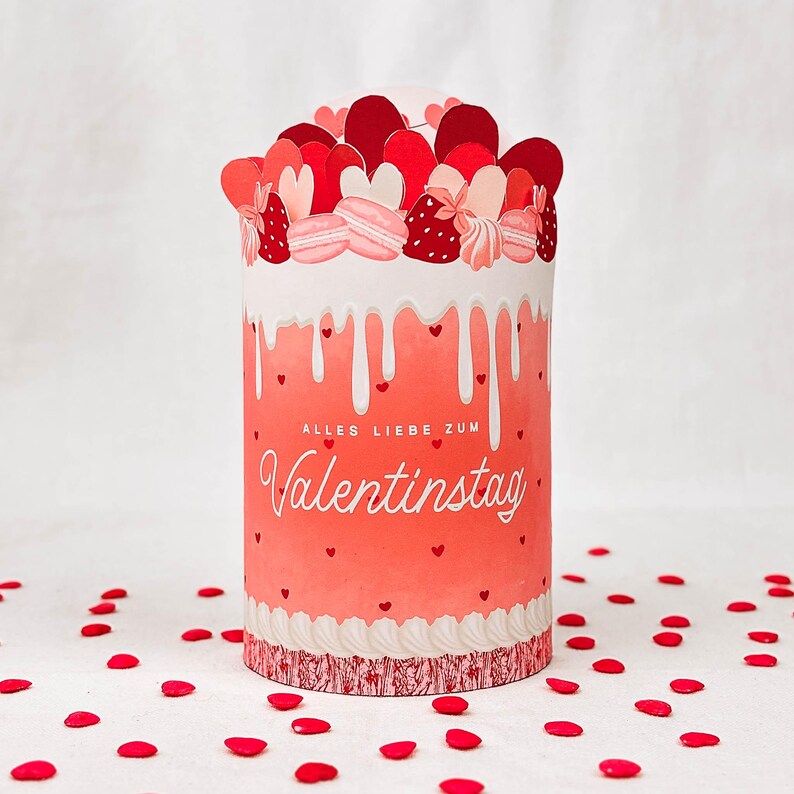 May include: A cylindrical Valentine's Day gift box with a red and pink design. The box features a dripping white frosting effect, heart accents, and the text "ALLES LIEBE ZUM Valentinstag". The top is decorated with heart-shaped cutouts and macarons.