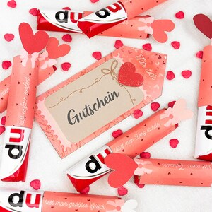 May include: A collection of wrapped chocolate bars with red and white packaging, decorated with heart-shaped cutouts and gift tags. The tags and wrappers feature German text and small heart-shaped confetti.