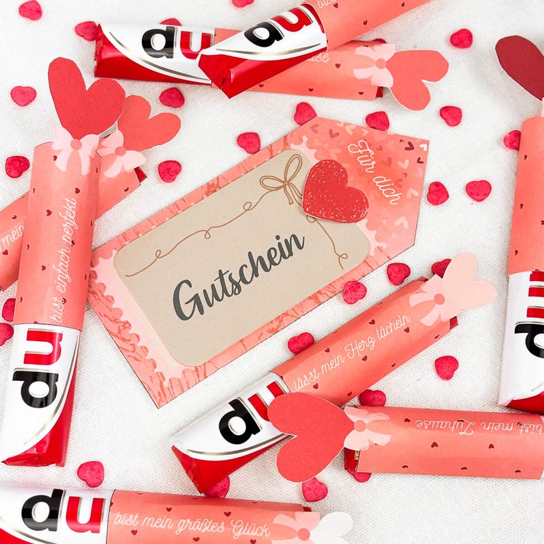 May include: A collection of wrapped chocolate bars with red and white packaging, decorated with heart-shaped cutouts and gift tags. The tags and wrappers feature German text and small heart-shaped confetti.