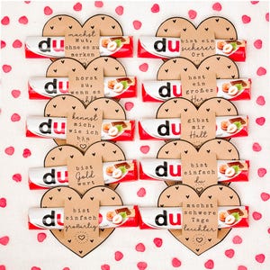 May include: Arrangement of chocolate bars with the word "du" on the wrapper, arranged in heart shapes with German phrases on brown paper. Red and white wrappers with hazelnut chocolate. Scattered red heart-shaped confetti.