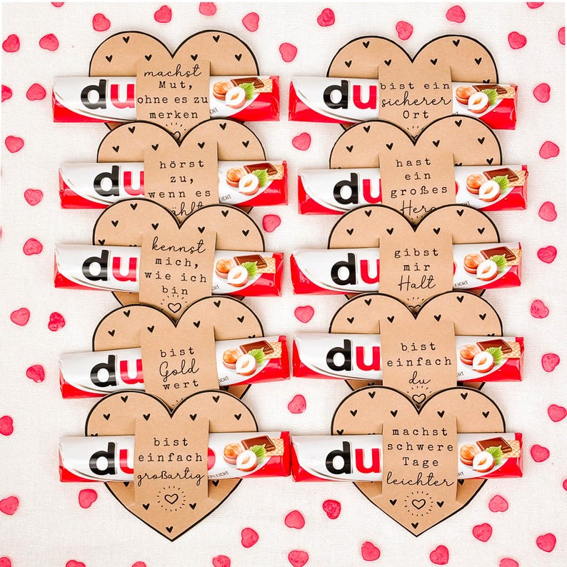 May include: Arrangement of chocolate bars with the word "du" on the wrapper, arranged in heart shapes with German phrases on brown paper. Red and white wrappers with hazelnut chocolate. Scattered red heart-shaped confetti.