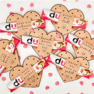 May include: A collection of heart-shaped cards with German text, each paired with a chocolate bar. The cards are brown with black heart accents and German phrases. Pink heart confetti is scattered around the arrangement.