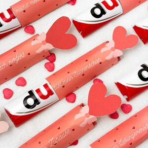 May include: Several Kinder Bueno chocolate bars wrapped in pink paper with heart-shaped accents and text. Each wrapper has a red heart attached with a bow. Scattered around are small red heart confetti pieces. The text on the wrappers is in German.