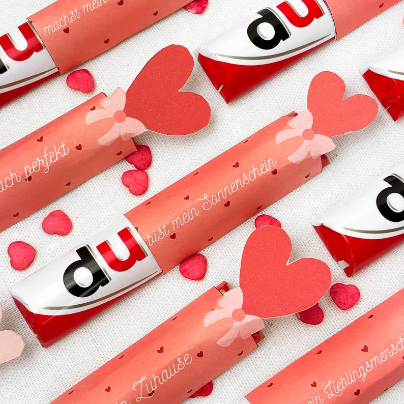 May include: Several Kinder Bueno chocolate bars wrapped in pink paper with heart-shaped accents and text. Each wrapper has a red heart attached with a bow. Scattered around are small red heart confetti pieces. The text on the wrappers is in German.