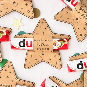 May include: Several star-shaped kraft paper holders featuring black star and sparkle illustrations, each securing a red and white chocolate bar. The center holder displays the text 'bist ein heller Stern' with images of hazelnuts and chocolate.