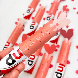 May include: A collection of pink and white candy bars with red accents and the word "du" printed on them. The candy bars are wrapped in pink paper with the text "l&auml;sst mein Herz l&auml;cheln" and small red hearts. A hand holds one of the candy bars.
