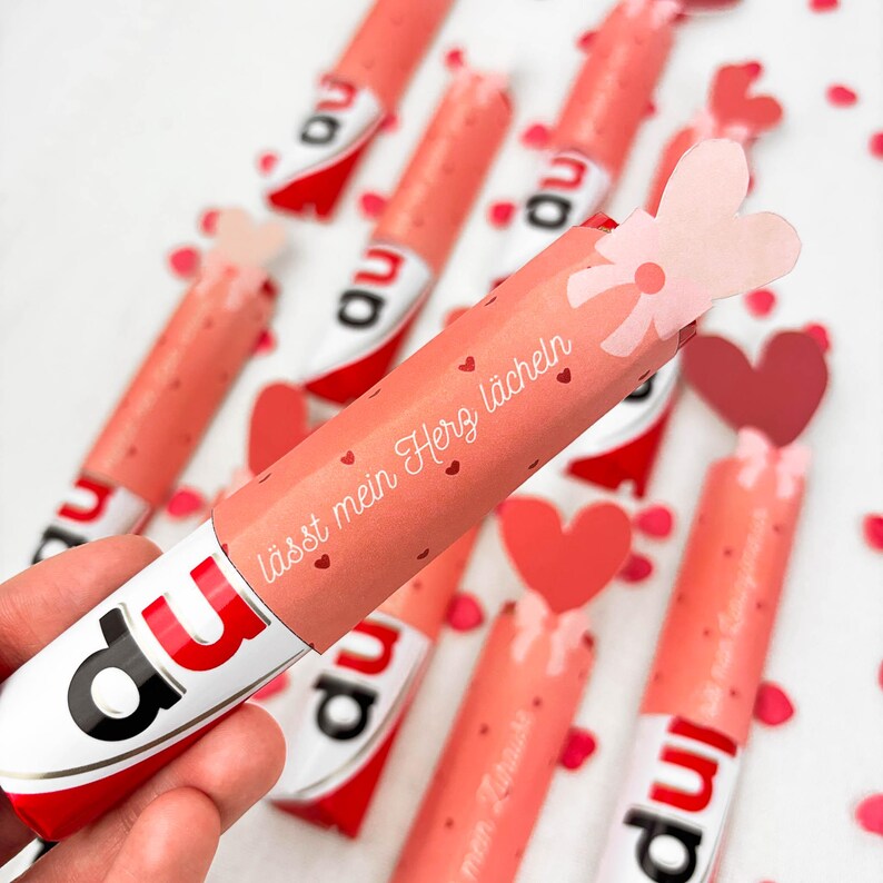May include: A collection of pink and white candy bars with red accents and the word "du" printed on them. The candy bars are wrapped in pink paper with the text "l&auml;sst mein Herz l&auml;cheln" and small red hearts. A hand holds one of the candy bars.
