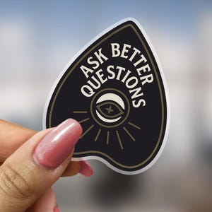 May include: A black and gold sticker in the shape of a planchette, held by a hand with pink nails. The sticker has the text "ASK BETTER QUESTIONS" in white, with an eye symbol in the center.