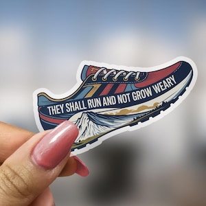 May include: A sticker in the shape of a running shoe, featuring a mountain landscape and the text "THEY SHALL RUN AND NOT GROW WEARY." The shoe design incorporates red, blue, and gold colors, with white laces and a white sole. The sticker is held up by a hand.