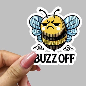 May include: A white sticker with a cartoon bee with a grumpy face and blue wings. The bee is flying above a cloud and the text "Buzz Off" is below the bee.