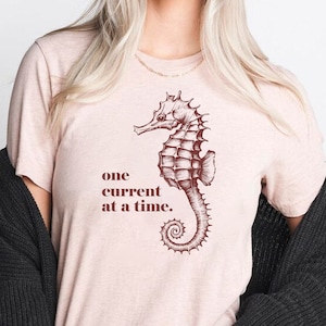 May include: A light pink t-shirt featuring a detailed brown seahorse illustration and the text "one current at a time." The shirt is made of a soft material and is a casual wear item.