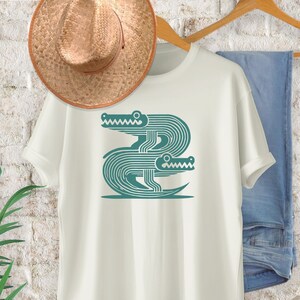 Peut inclure: T-shirt blanc avec un graphique turquoise de deux alligators stylis&eacute;s entrelac&eacute;s.