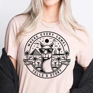 May include: A pink t-shirt with a black graphic of a camel wearing sunglasses in front of a desert scene with pyramids and palm trees. The graphic says "Where Every Camel Tells A Story".