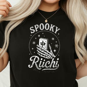 May include: Black t-shirt with a distressed white graphic. The graphic features the text "Spooky Riichi" with a skeleton hand holding a mahjong tile. The design is surrounded by stars and a circle.