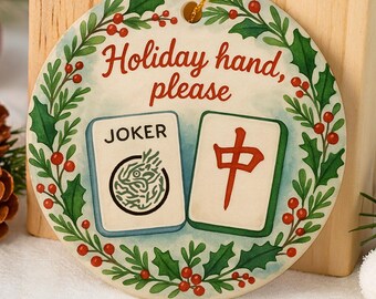 May include: Round ceramic holiday ornament with the text "Holiday hand, please." The ornament features two Mahjong tiles and is surrounded by a green and red holly wreath. The background includes a wooden block, red ornaments, and pine cones.