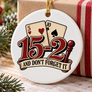 May include: A round white ceramic ornament featuring a graphic of a five of hearts and a ten of spades playing card. The text 15-2 AND DON'T FORGET IT is printed in red and black with a cribbage peg illustration on a gold hanging string.