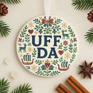 May include: Round ceramic ornament with the text "UFF DA" in navy blue, surrounded by a floral and Viking ship design in red, green, and gold. A white ribbon is attached for hanging. The ornament is decorated with snowflakes, hearts, and deer.