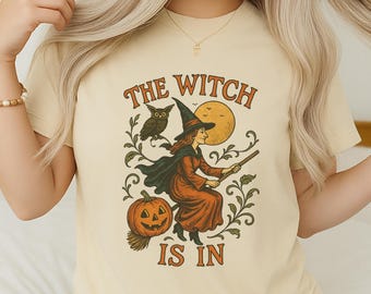 May include: Beige t-shirt with a Halloween-themed graphic. The design features a witch on a broom, an owl, a pumpkin, and the text "THE WITCH IS IN" in orange and brown. The witch is wearing a black hat and orange dress.
