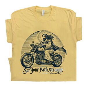Puede incluir: Una camiseta amarilla con una ilustraci&oacute;n en blanco y negro de Jes&uacute;s conduciendo una motocicleta. El texto "Set Your Path Straight - (Proverbs 3:6)" est&aacute; impreso debajo de la imagen.