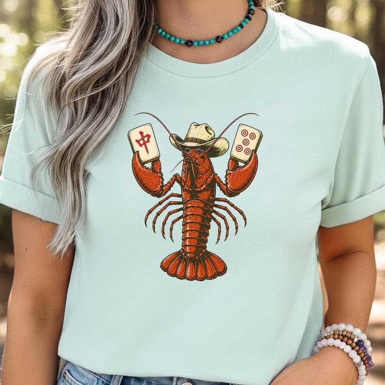May include: A light mint green t-shirt featuring a graphic illustration of a red lobster wearing a tan cowboy hat. The lobster is holding a mahjong tile with a red Chinese character in its left claw and a mahjong tile with five red circles in its right.