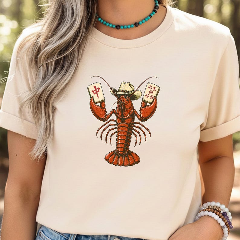 May include: A cream-colored t-shirt featuring a graphic of a red lobster wearing a cowboy hat and holding two mahjong tiles. The lobster is centered on the chest, with one tile displaying the Chinese character for middle and the other five dots.