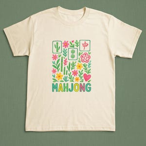 May include: Cream-colored t-shirt featuring a colorful floral design with the word "MAHJONG" in green, yellow, and blue. The design includes bamboo, flowers, and a heart, creating a playful and vibrant aesthetic.