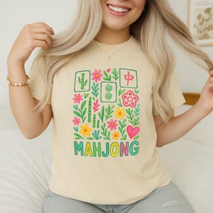 May include: A light beige t-shirt featuring a colorful Mahjong-themed graphic. The design includes bamboo, flowers, and the word "MAHJONG" in green. The shirt is a casual, everyday wear item.