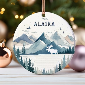 May include: A round ceramic ornament with a watercolor painting of a mountain range, a lake, and a moose. The text "ALASKA" is written in the center of the ornament.