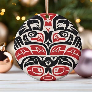 May include: Round ceramic ornament with a red, black, and white design. The design features a stylized animal face with geometric patterns.