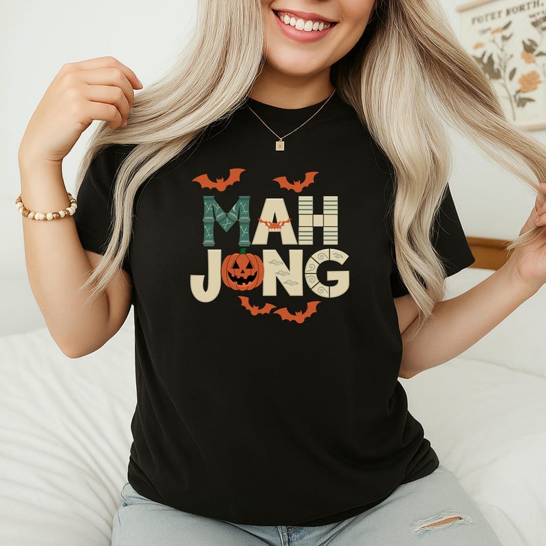 May include: Black t-shirt with a Halloween-themed "MAH JONG" graphic. The design includes bats, a jack-o'-lantern, and the game's name in a playful font. The shirt is a comfortable, casual style.