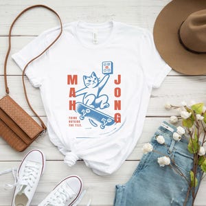 May include: White t-shirt with a blue and red graphic of a cat skateboarding with the word "Mahjong" written vertically on the left and right sides of the graphic. The text "Think outside the tile." is written below the graphic.