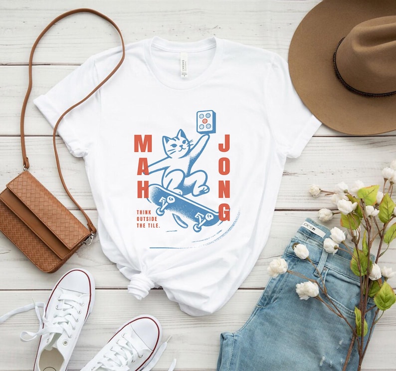 May include: White t-shirt with a blue and red graphic of a cat skateboarding with the word "Mahjong" written vertically on the left and right sides of the graphic. The text "Think outside the tile." is written below the graphic.