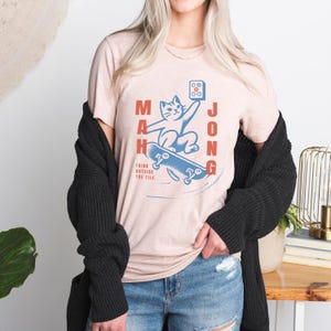 May include: A light pink t-shirt with a blue and red graphic of a cat riding a skateboard. The graphic says "Mah Jong" and "Think outside the tile."