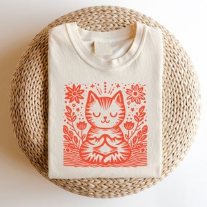 以下が含まれることがあります： クリーム色のTシャツに、赤い花の模様の中に瞑想している猫のイラストが描かれています。