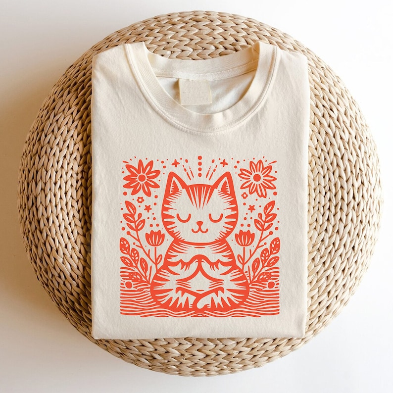 以下が含まれることがあります： クリーム色のTシャツに、赤い花の模様の中に瞑想している猫のイラストが描かれています。
